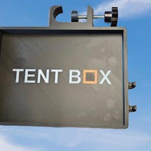 May include: A dark grey rectangular device with the text "TENT BOX" in white. The letter "O" is a small orange square. The device has a clamp, a screw, and two small side attachments. The background is a blue sky.