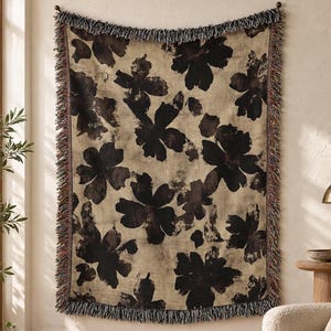 May include: A rectangular woven throw blanket featuring a large-scale dark brown floral pattern on a beige background. The blanket has a decorative fringe border along all four sides with multi-colored threads woven into the edges.