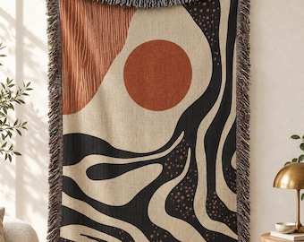 May include: A woven throw blanket featuring an abstract design with a large terracotta circle, wavy black lines, and textured beige sections. The blanket has a fringed edge along the top and bottom, displaying a multi-colored thread pattern.
