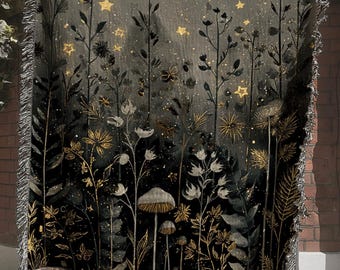 May include: A woven throw blanket featuring a dark, nocturnal forest scene with golden stars, a crescent moon, tall wildflowers, and mushrooms. The design transitions from a deep black base to a lighter gray top, finished with a fringed edge.
