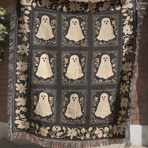 May include: A black and cream woven throw blanket featuring a grid of nine rectangular panels, each containing a single ghost illustration. The blanket is bordered by a floral vine pattern and finished with a fringed edge on all four sides.