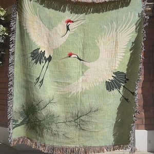 May include: A woven throw blanket featuring a light green background with two large white cranes in flight. Each crane has a red crown and black tail feathers. A dark green pine branch is depicted at the bottom. The blanket has a fringed edge.