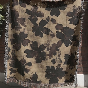 May include: A rectangular woven throw blanket featuring a repeating dark brown floral pattern on a tan background. The blanket has a textured appearance with frayed, multi-colored fringe edges along all four sides, creating a soft, decorative finish.