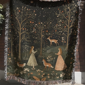 May include: A woven tapestry throw blanket featuring a forest scene with trees, deer, and figures in long dresses. The blanket has a dark green background with a fringed edge. The artwork includes shades of brown, orange, and cream.