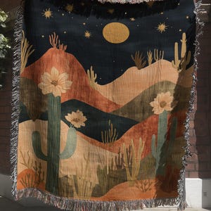 May include: A woven throw blanket featuring a desert landscape at night with a large yellow moon, stars, and blooming cacti. The design includes rolling hills in shades of orange, brown, and dark blue, finished with a decorative fringed edge.
