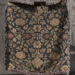 May include: A woven throw blanket featuring a dark charcoal background with a repeating floral pattern in shades of muted orange, tan, and cream. The rectangular textile has a decorative fringe along all four edges and a symmetrical design.