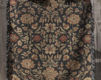 May include: A woven throw blanket featuring a dark charcoal background with a repeating floral pattern in shades of muted orange, tan, and cream. The rectangular textile has a decorative fringe along all four edges and a symmetrical design.