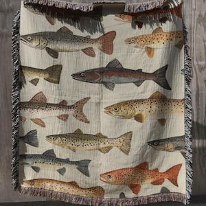 May include: A woven throw blanket featuring a repeating pattern of various freshwater fish species in shades of brown, tan, and grey. The blanket has a cream-colored background and is finished with decorative fringed edges on the top and bottom.