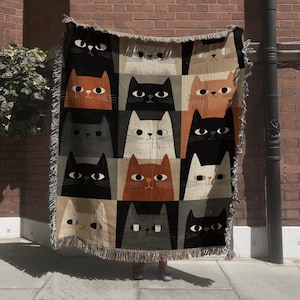 May include: A woven throw blanket featuring a repeating pattern of stylized cat faces in black, orange, gray, and white. The blanket has a fringed edge and is displayed against a brick wall.