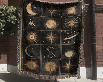 May include: A rectangular woven throw blanket featuring a grid of twelve celestial illustrations in gold and cream on a black background. The design includes various phases of the moon, suns, stars, and planetary rings, finished with a fringed edge.