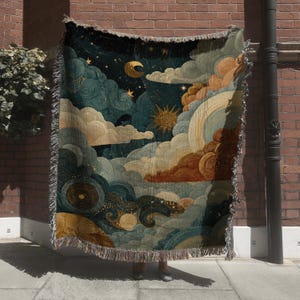 May include: A woven throw blanket featuring a celestial design with a night sky, clouds, a sun, and a crescent moon in shades of teal, gold, and brown. The blanket has a fringed edge.