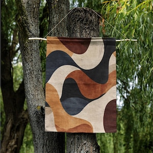 Pu&ograve; includere: Un arazzo da parete rettangolare con un motivo astratto a onde nei toni del marrone, beige, nero e blu navy. L'opera &egrave; sospesa a un'asta di legno, davanti a un albero e un fogliame verde.
