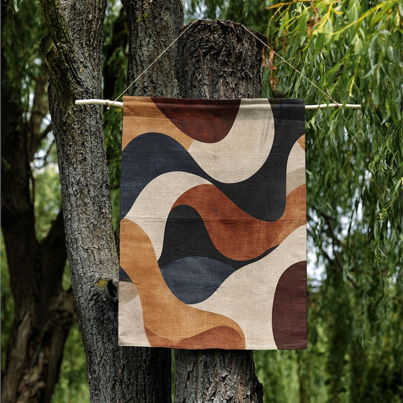 Pu&ograve; includere: Un arazzo da parete rettangolare con un motivo astratto a onde nei toni del marrone, beige, nero e blu navy. L'opera &egrave; sospesa a un'asta di legno, davanti a un albero e un fogliame verde.