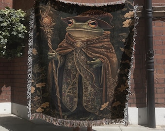 May include: A woven throw blanket featuring a detailed illustration of a green frog dressed as a wizard in a brown cloak and hat, holding a staff. The design includes floral patterns in shades of brown, gold, and black with a fringed edge.