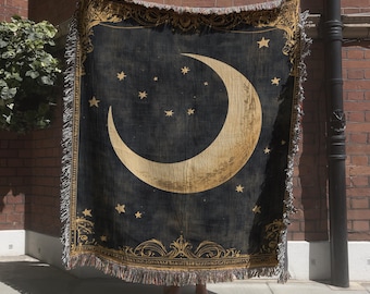Peut inclure: Un plaid tissé rectangulaire présentant un grand croissant de lune doré et des étoiles éparpillées sur un fond noir profond. La couverture est encadrée par une bordure dorée ornementale et finie avec des franges sur les quatre côtés.