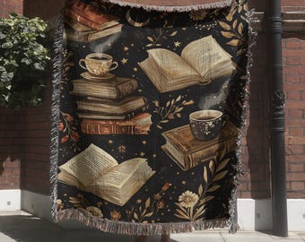 May include: A black woven throw blanket featuring a repeating pattern of open and stacked books, steaming cups of coffee, crescent moons, stars, and botanical floral elements. The blanket has a fringed edge and a dark, moody color palette.