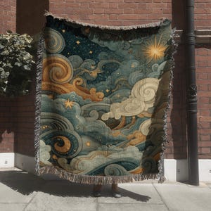 May include: A woven throw blanket featuring a celestial design with swirling clouds, stars, and a bright sun. The color palette includes shades of blue, gold, and cream. The blanket has a fringed edge and is displayed outdoors.