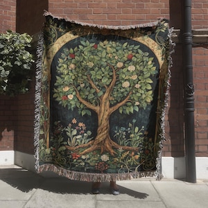May include: A woven tapestry throw blanket featuring a detailed tree design with leaves, flowers, and roots. The color palette includes shades of green, brown, and cream, with a dark blue background. The blanket has a fringed edge.