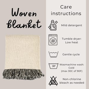 Op de afbeelding: Een beige geweven deken met zwarte en witte franjes. Aan de rechterkant van de afbeelding staan ​​wasinstructies, waaronder instructies voor wassen, drogen en bleken.