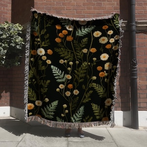 May include: A black throw blanket with a floral pattern featuring white and orange daisies and green foliage. The blanket has a fringed edge and is displayed against a brick wall.