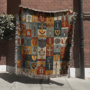 May include: A woven throw blanket featuring a repeating pattern of medieval shields, swords, and crosses in shades of red, blue, yellow, and beige. The blanket has a fringed edge and is displayed against a brick wall.