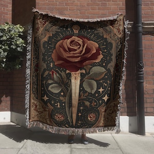 May include: A woven tapestry blanket featuring a large rose pierced by a sword. The design is in shades of burgundy, gold, and green on a dark background. The blanket has a fringed edge and a decorative border.