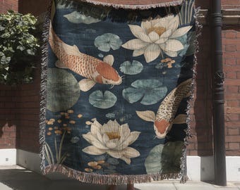 May include: A woven throw blanket featuring a dark blue background with two koi fish in orange and white, surrounded by large white lotus flowers and green lily pads. The blanket has a decorative fringed edge along the top and bottom borders.