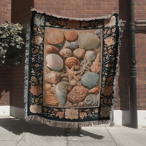 May include: A rectangular woven throw blanket featuring a central panel of various seashells and a starfish in shades of orange, blue, and tan. The blanket has a dark border decorated with floral patterns and a fringed edge on all four sides.