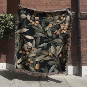 May include: A dark-colored throw blanket featuring a botanical olive branch design. The blanket has a black background with olive branches in shades of green, brown, and tan. The edges are finished with a fringe.