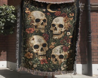 May include: A rectangular woven throw blanket featuring a dark background with four human skulls surrounded by colorful flowers and foliage. A golden crescent moon is positioned at the top center. The blanket has a fringed edge on all four sides.