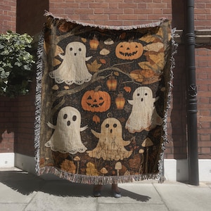 May include: A woven Halloween-themed throw blanket featuring a dark background with illustrations of ghosts, jack-o'-lanterns, and autumn leaves. The blanket has a fringed edge and is held up against a brick wall.
