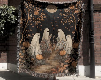 May include: A woven throw blanket featuring a dark background with four white ghost figures standing among orange pumpkins and floral patterns. The design includes a large pale moon at the top, stylized trees, and a fringed edge on all sides.