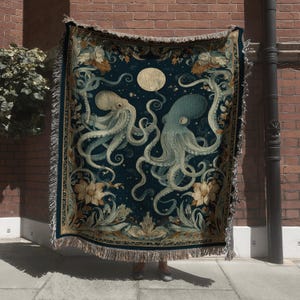 May include: A dark blue woven throw blanket featuring two octopuses with swirling tentacles, a full moon, and floral accents. The blanket has a fringed edge and a detailed, vintage-inspired design. The color palette includes shades of blue, cream, and gold.