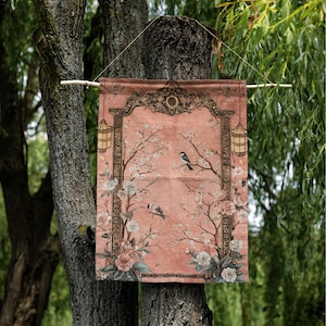May include: A decorative wall hanging with a pink background, featuring a floral and bird design. The artwork includes blossoming branches, birds, and ornate framing with lanterns. The hanging is suspended from a wooden rod.
