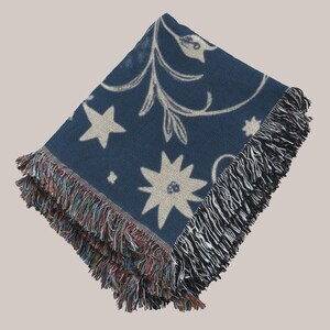 Op de afbeelding: Een blauwe geweven deken met een wit bloemenpatroon en sterdesigns. De deken heeft een franje rand.