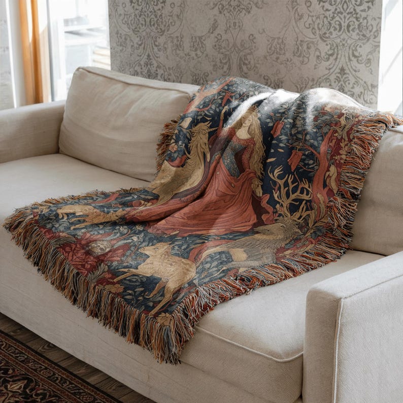 Pu&ograve; includere: Una coperta tessuta con un design dettagliato in stile arazzo, raffigurante una figura centrale in abito rosso, un unicorno e vari animali su uno sfondo floreale blu scuro, rifinita con frange decorative lungo i bordi.