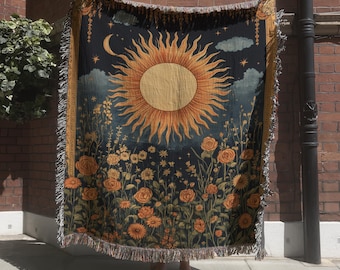 May include: A woven throw blanket featuring a large central sun, a crescent moon, and stars against a dark background, with a field of orange and yellow flowers at the bottom. The blanket has a fringed edge and a decorative border on both sides.