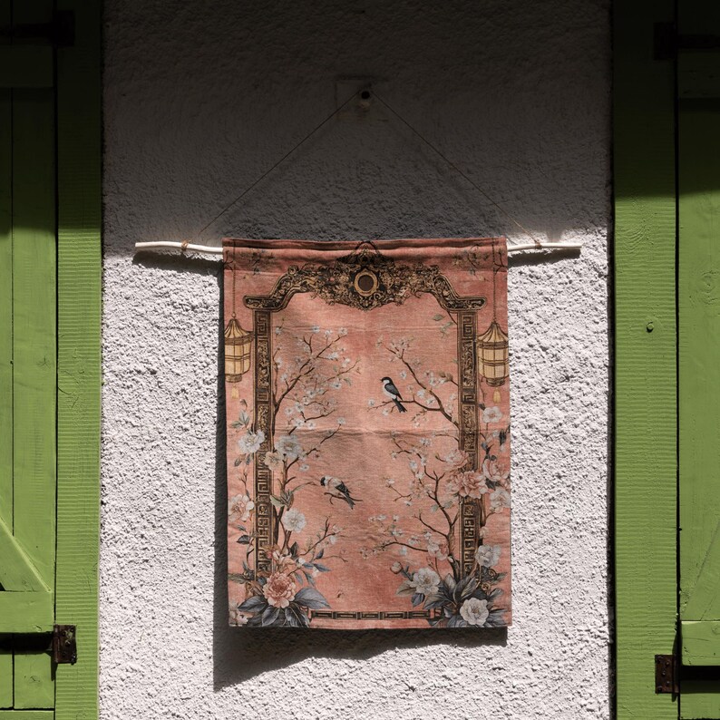May include: A rectangular, peach-colored wall hanging with a floral and bird design. The artwork features a decorative border, flowering branches, and two birds. The hanging is suspended from a wooden rod.
