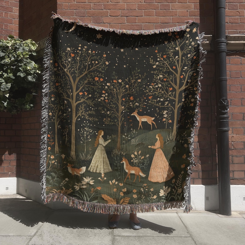 May include: A woven tapestry throw blanket featuring a forest scene with trees, deer, and figures in long dresses. The blanket has a dark green background with a fringed edge. The artwork includes shades of brown, orange, and cream.