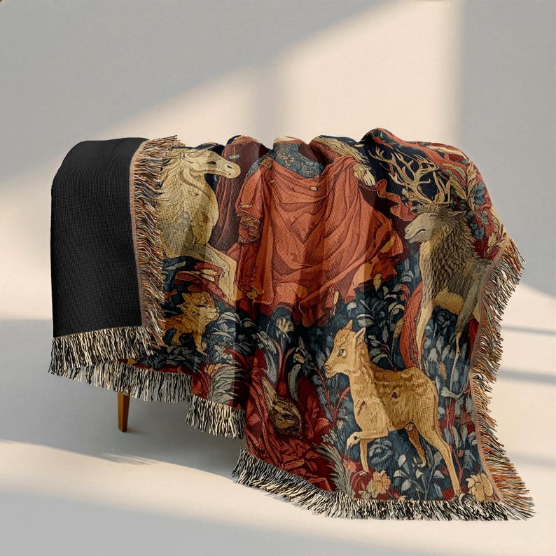 Pu&ograve; includere: Una coperta tessuta con un design dettagliato in stile arazzo che raffigura un unicorno, un cervo e un piccolo cerbiatto tra motivi floreali blu scuro e rossi. La coperta ha un retro nero a tinta unita e frange decorative sui bordi.