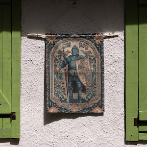 K&ouml;nnte beinhalten: Ein Wandbehang mit einem Ritter in voller R&uuml;stung, der ein Schwert und einen Schild h&auml;lt. Der Wandbehang hat einen detaillierten floralen Rand in Blau-, Beige- und Gr&uuml;nt&ouml;nen. Er h&auml;ngt an einer wei&szlig;en Stuckwand mit gr&uuml;nen Fensterl&auml;den.