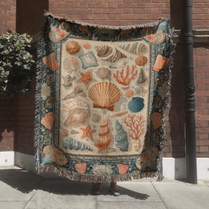 May include: A beige throw blanket featuring a variety of seashells, starfish, and coral in shades of orange, blue, and brown. The blanket has a decorative border and fringe.