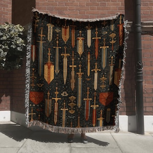 May include: A dark woven throw blanket featuring a repeating pattern of swords in shades of gold, red, and brown against a black background. The blanket has a fringed edge and is held up against a brick wall.