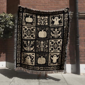 May include: Black and beige Halloween-themed throw blanket. The blanket features a repeating pattern of pumpkins, ghosts, and floral designs within square blocks. The edges are finished with a fringe.