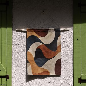 Pu&ograve; includere: Un arazzo da parete rettangolare con un motivo ondulato nei toni del marrone, beige, blu navy e bianco. Il tessuto &egrave; sospeso a un'asta di legno contro un muro bianco strutturato. Il pezzo &egrave; esposto vicino a un'imposta verde.