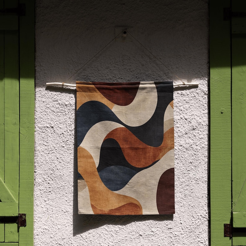 Pu&ograve; includere: Un arazzo da parete rettangolare con un motivo ondulato nei toni del marrone, beige, blu navy e bianco. Il tessuto &egrave; sospeso a un'asta di legno contro un muro bianco strutturato. Il pezzo &egrave; esposto vicino a un'imposta verde.