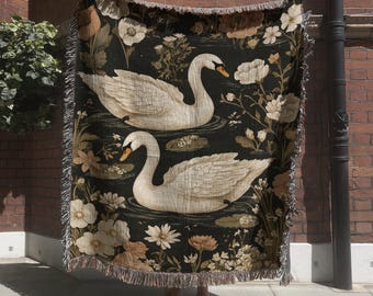 May include: A woven throw blanket featuring a dark background with two white swans swimming among various light-colored flowers and foliage. The blanket has a textured appearance with a fringed edge along the top and bottom borders.