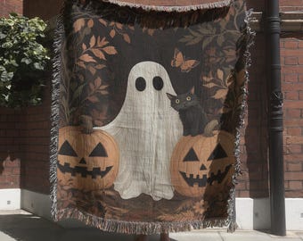 May include: A woven throw blanket featuring a central white ghost figure flanked by two orange jack-o'-lanterns and a black cat. The background displays dark brown autumn foliage and a small butterfly. The blanket has a fringed edge on all sides.