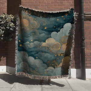 May include: A woven throw blanket featuring a celestial design with teal, gold, and cream colors. The blanket depicts clouds, stars, and swirling patterns. The edges have a fringed trim.