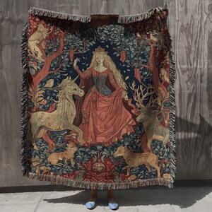 Pu&ograve; includere: Una coperta in tessuto arazzo raffigurante una figura centrale con lunghi capelli biondi in un abito rosso e blu, circondata da un unicorno, un leone e altri animali in un ambiente boschivo. La coperta rettangolare ha un bordo decorativo a frange.